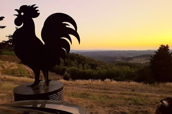 Silhouette of a rooster statue at sunset over a hilly landscape.