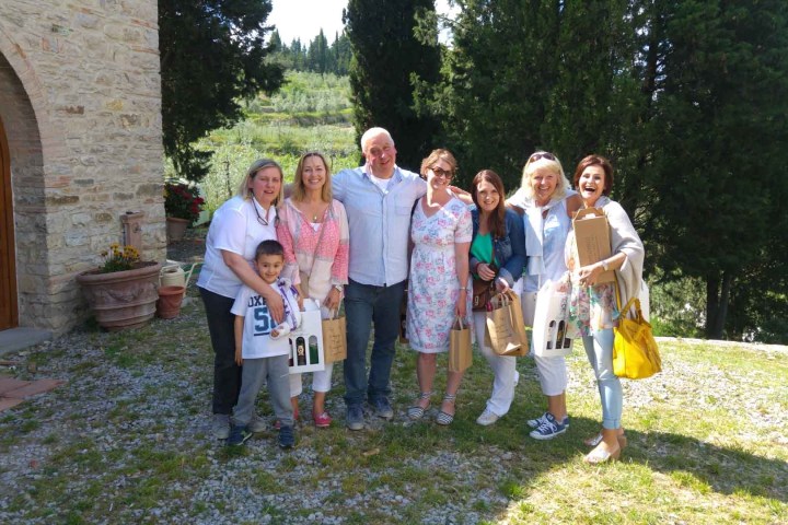 Group of people smiling outdoors, holding bags, near a stone building and trees.