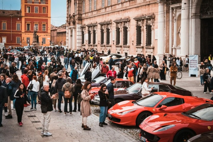 Crowded street with people viewing red sports cars in front of historic buildings.
