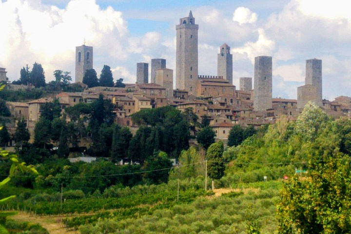 Medieval town with tall stone towers and lush greenery in the foreground under a cloudy sky.