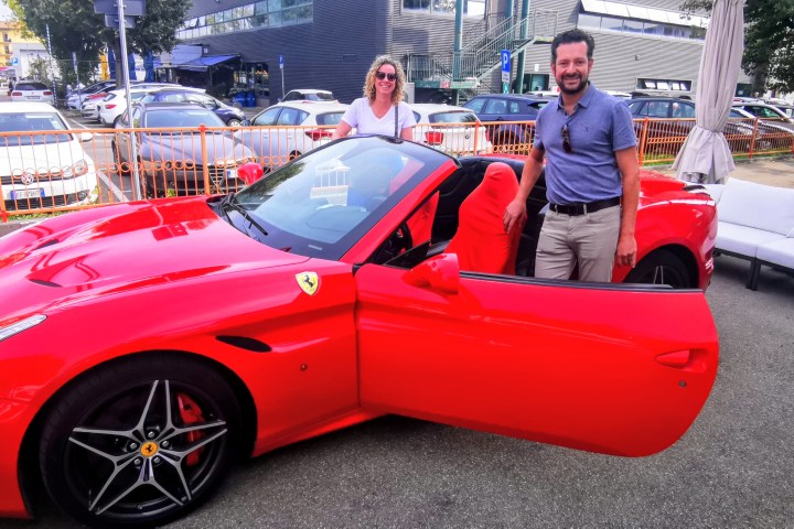 Two people stand by a red convertible sports car in a parking lot.
