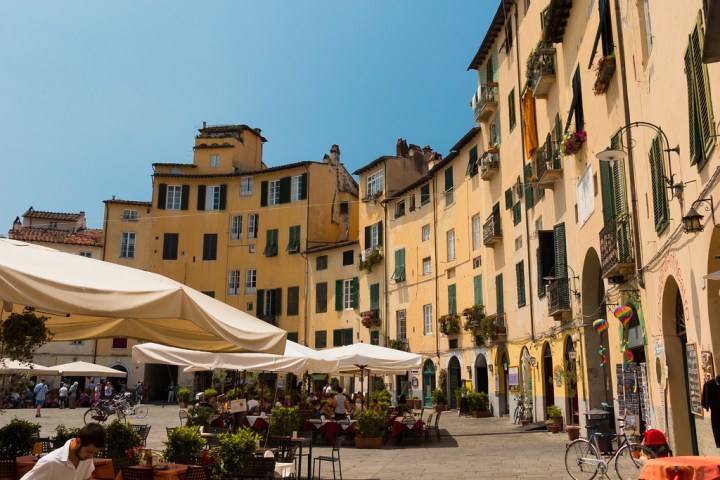 Outdoor cafe in an Italian square surrounded by yellow buildings with green shutters.