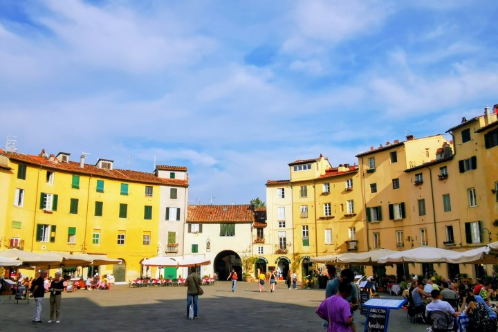Outdoor square with people dining under umbrellas, surrounded by yellow buildings on a sunny day.