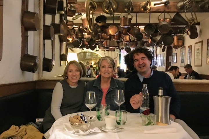 Three people sit at a restaurant table with wine glasses, surrounded by hanging copper pots.