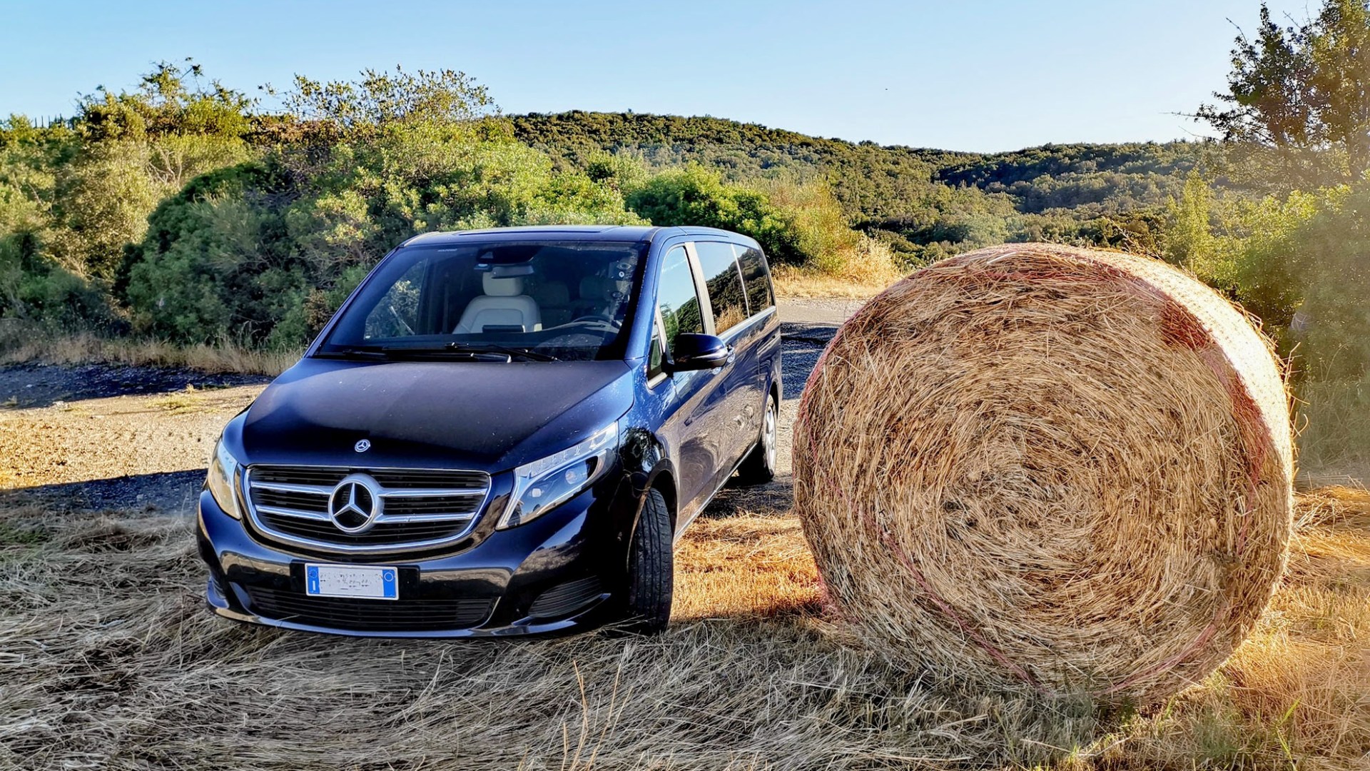 Black van parked next to large hay bale in a rural setting with trees in the background.